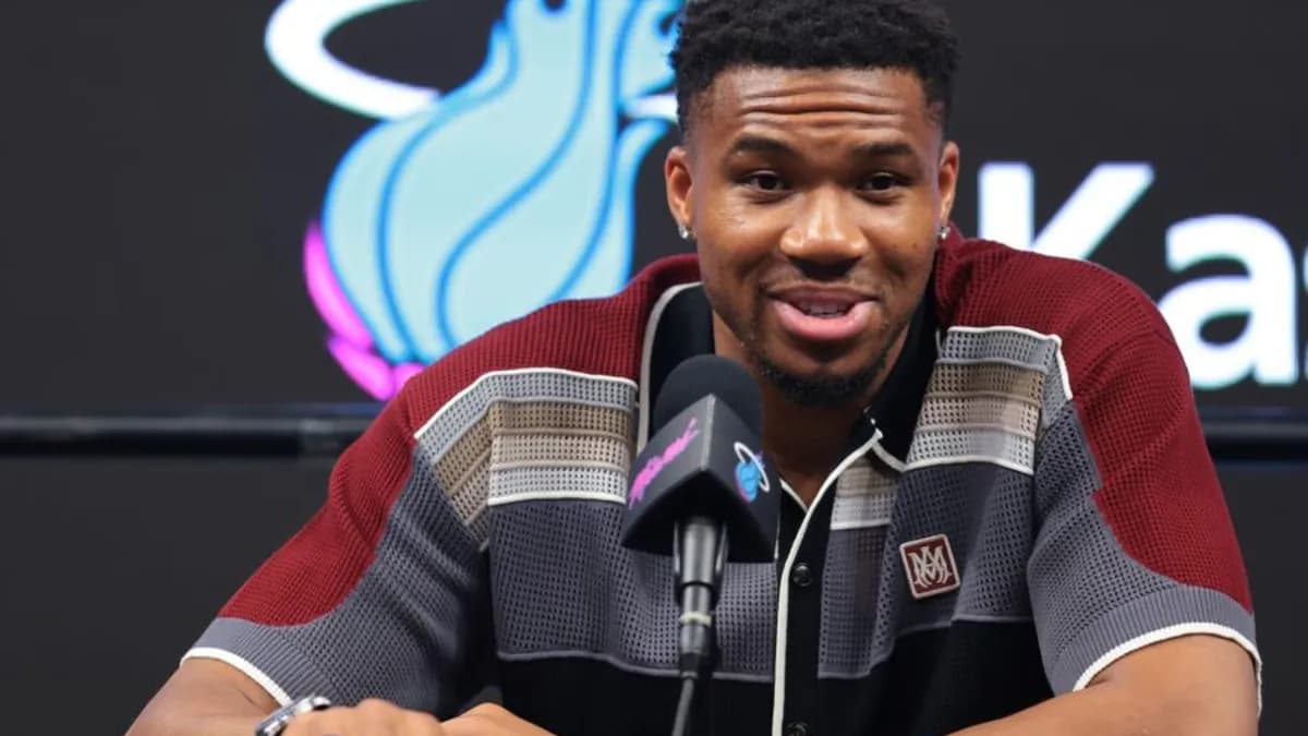 Giannis Antetokounmpo speaks into a microphone in front of a Miami Heat logo backdrop at a press conference