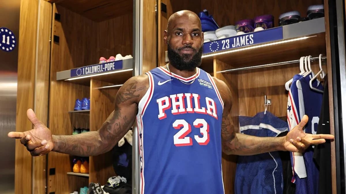 LeBron James poses with arms outstretched in a blue Philadelphia 76ers number 23 jersey in the team locker room