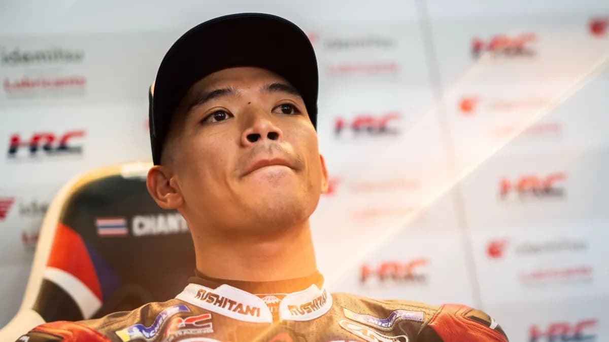 Somkiat Chantra in a black cap and Honda race suit sits in the garage in front of HRC branded boards and a seat bearing a Thai flag