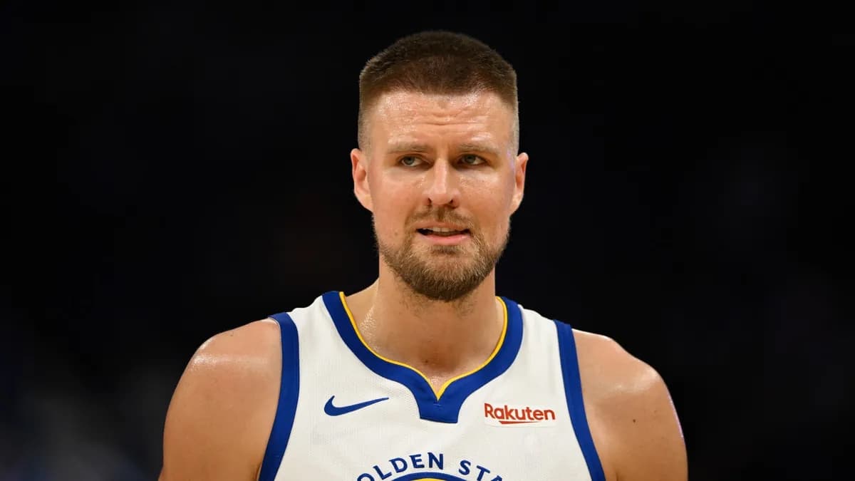 Kristaps Porzingis in a white Golden State Warriors jersey looks on during a game against a dark background