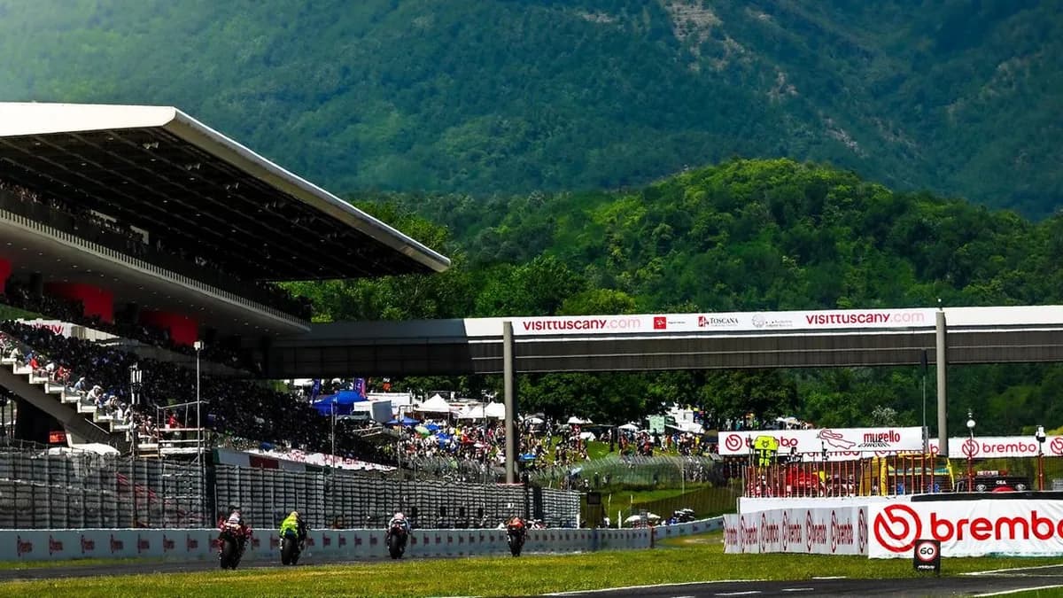 MotoGP riders race down Mugello’s main straight past the covered grandstand, with crowds and green Tuscan hills behind